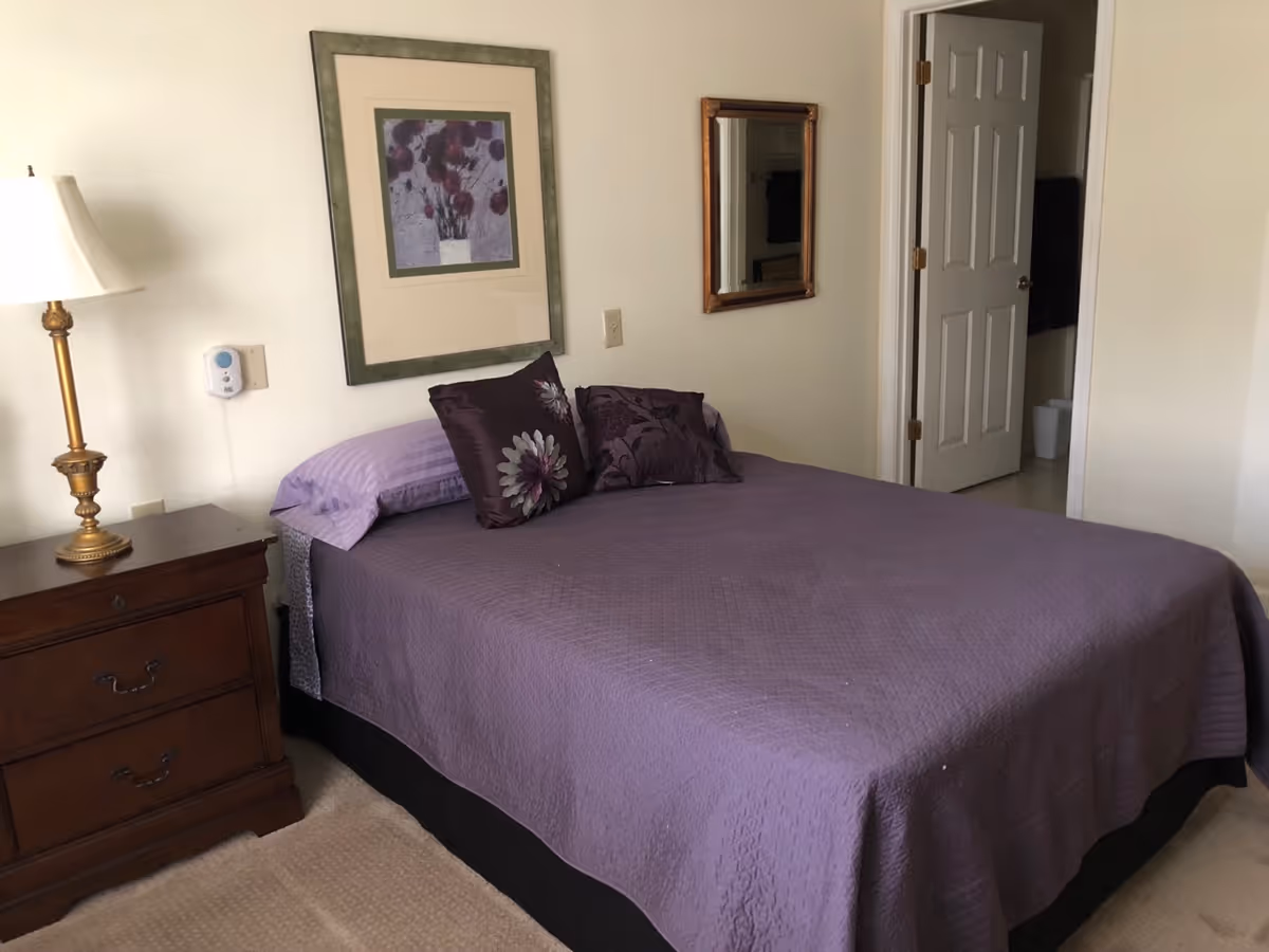 A bedroom with a bed covered in a purple quilt and two decorative pillows. There is a wooden nightstand with a lamp on the left side of the bed. A framed floral artwork hangs on the wall above the bed, and a mirror is mounted on the adjacent wall. An open door reveals a glimpse of a bathroom.