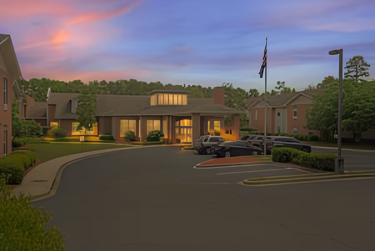 Exterior view of TerraBella Windsor Lake facility at dusk with a curved driveway, parked cars, an American flag on a flagpole, and surrounding greenery under a colorful sky.