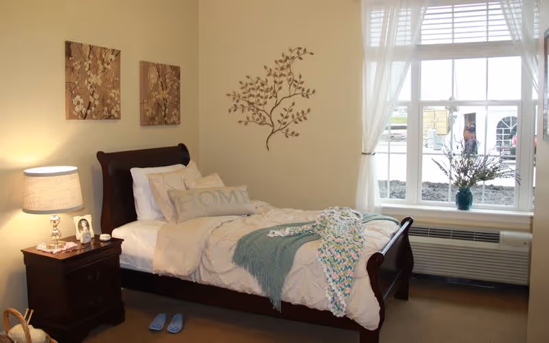 A cozy bedroom with a single dark wooden bed frame, white bedding, and decorative pillows including one with the word 'HOME'. A nightstand with a lamp, framed photo, and small items is beside the bed. Two floral paintings hang on the wall above the bed, and a metal wall art piece shaped like a leafy branch is on the adjacent wall. A large window with sheer white curtains lets in natural light, and a vase with flowers sits on the windowsill. Blue slippers are placed on the carpeted floor near the bed.