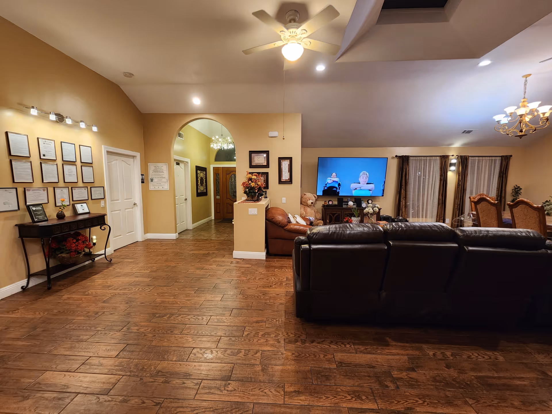 Interior view of a senior living facility common area with wooden flooring, a black leather sofa facing a wall-mounted TV showing two people, a brown leather couch, a large teddy bear, and a dining area with a chandelier and curtains in the background. The walls are painted beige with framed certificates and decorations, and there is a ceiling fan with a light.