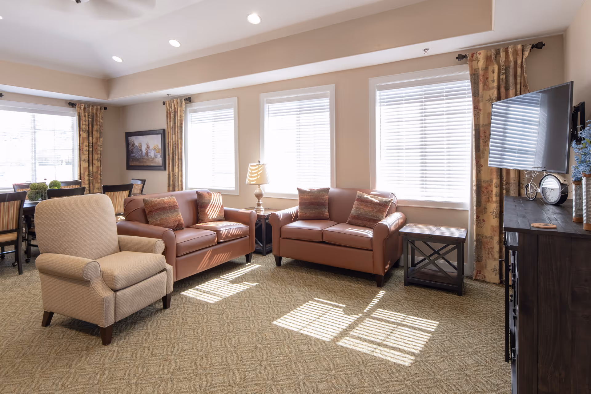 A bright and cozy living room with two brown leather sofas, a beige armchair, a wooden side table with a lamp, and a flat-screen TV mounted on a dark wooden cabinet. The room has large windows with white blinds and patterned curtains, and a dining area with a table and chairs is visible in the background.