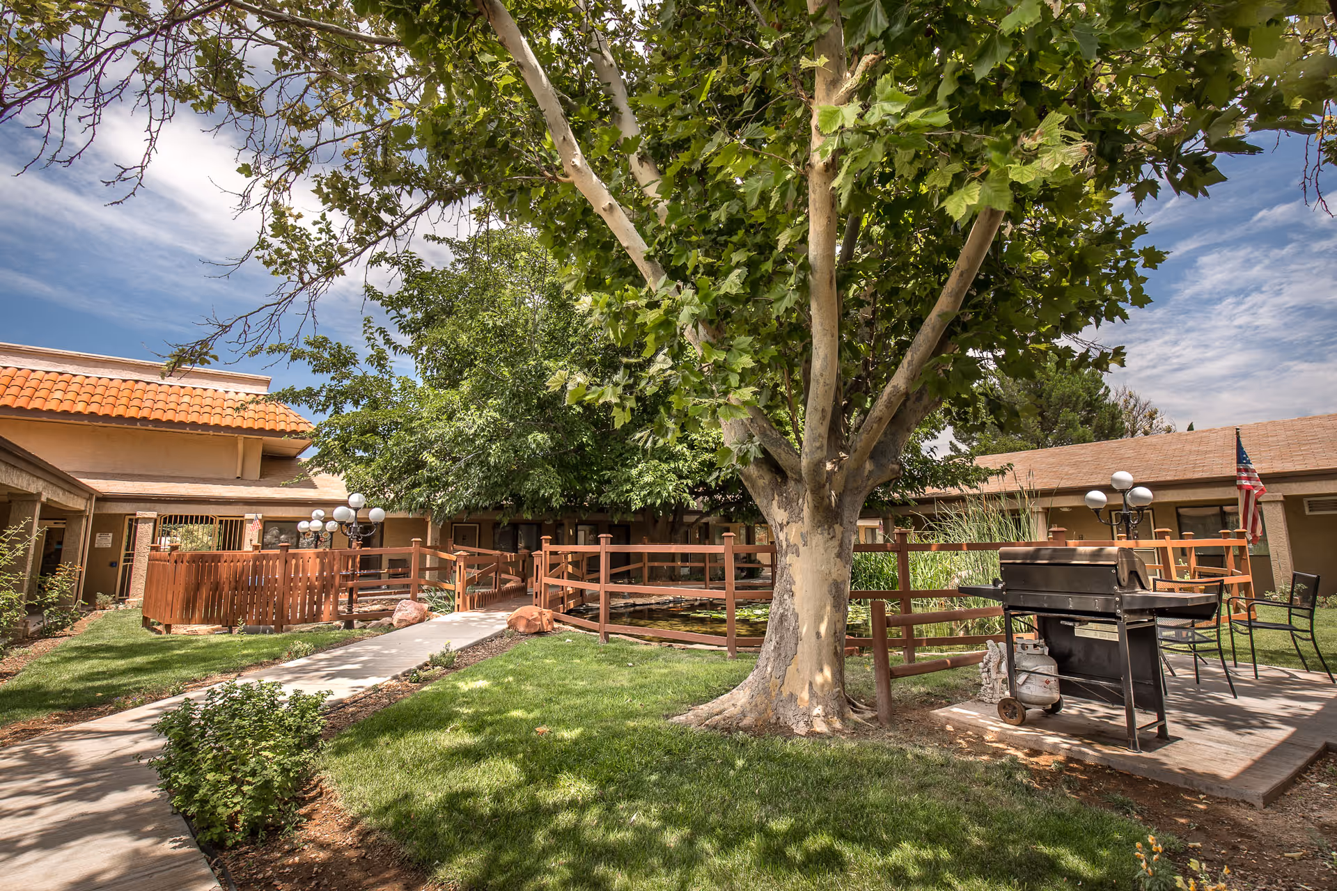 Outdoor courtyard area of Verde Valley Assisted Living featuring a large tree, green grass, a paved walkway, wooden railings, a barbecue grill on a concrete patio, and buildings with tiled roofs in the background under a partly cloudy sky.