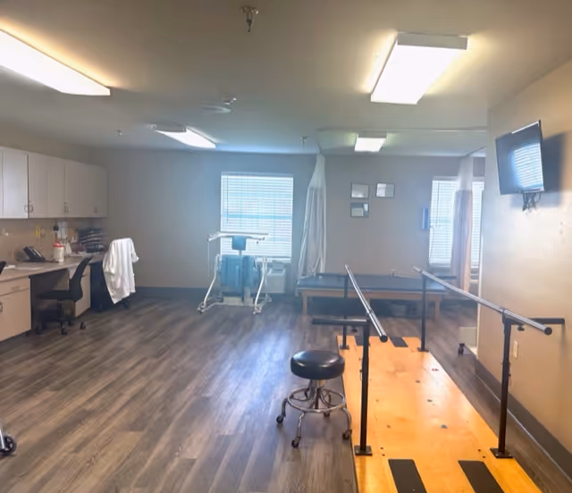 A rehabilitation or physical therapy room with wooden parallel bars for walking exercises, a stool on wheels, a treatment table, a walker, and a desk with a chair and medical supplies. The room has wood-look flooring, two windows with blinds, and a wall-mounted TV.