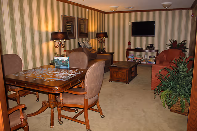 A cozy living room with striped wallpaper and beige carpet. The room features a wooden table with chairs and a partially completed jigsaw puzzle, two armchairs, a coffee table, a TV mounted on the wall, a shelf with board games and a coffee maker, two table lamps, framed pictures on the wall, and a large potted plant.