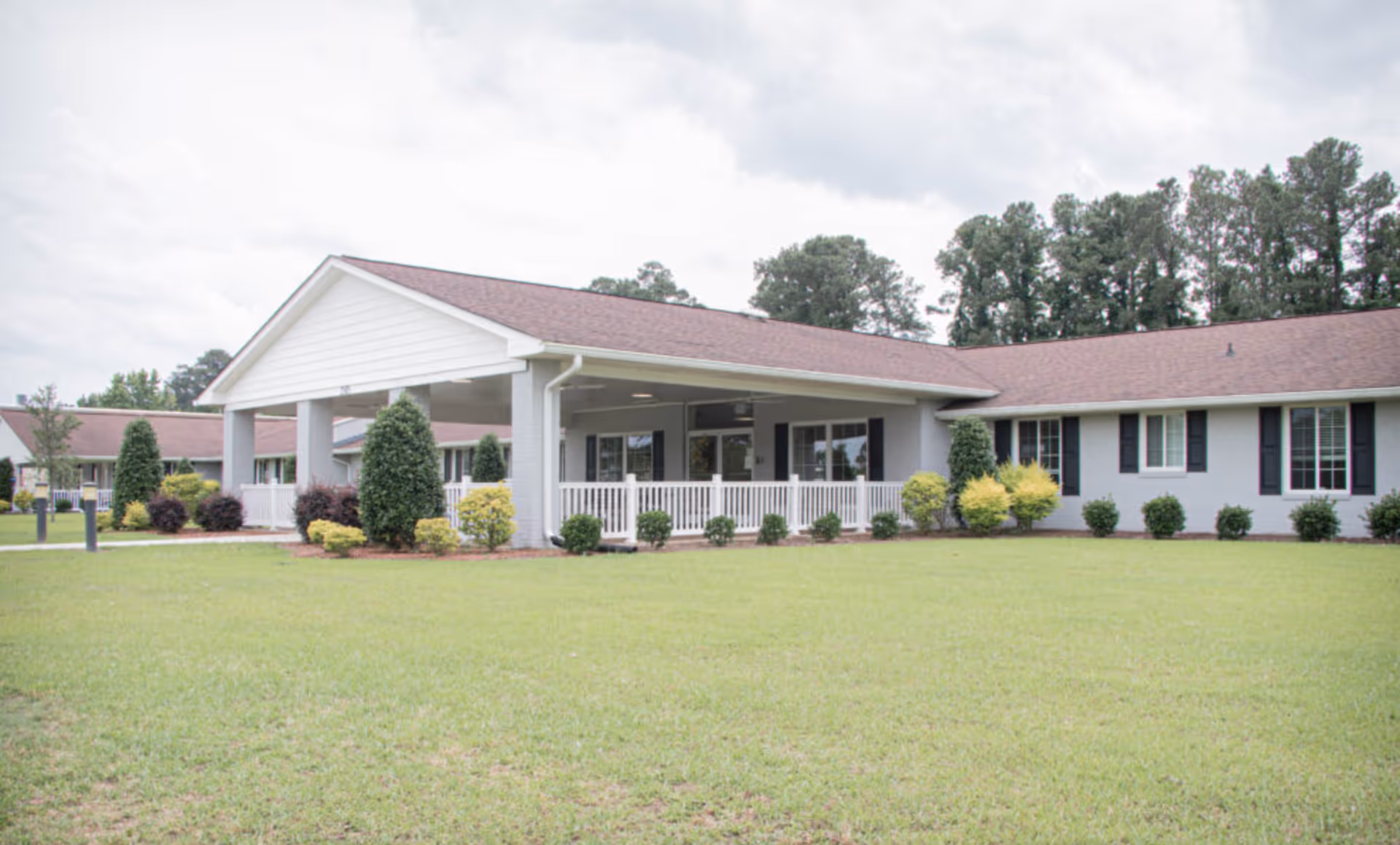 Single-story light-gray building with a covered entrance, white porch railing, shrubs, and a large grassy lawn.
