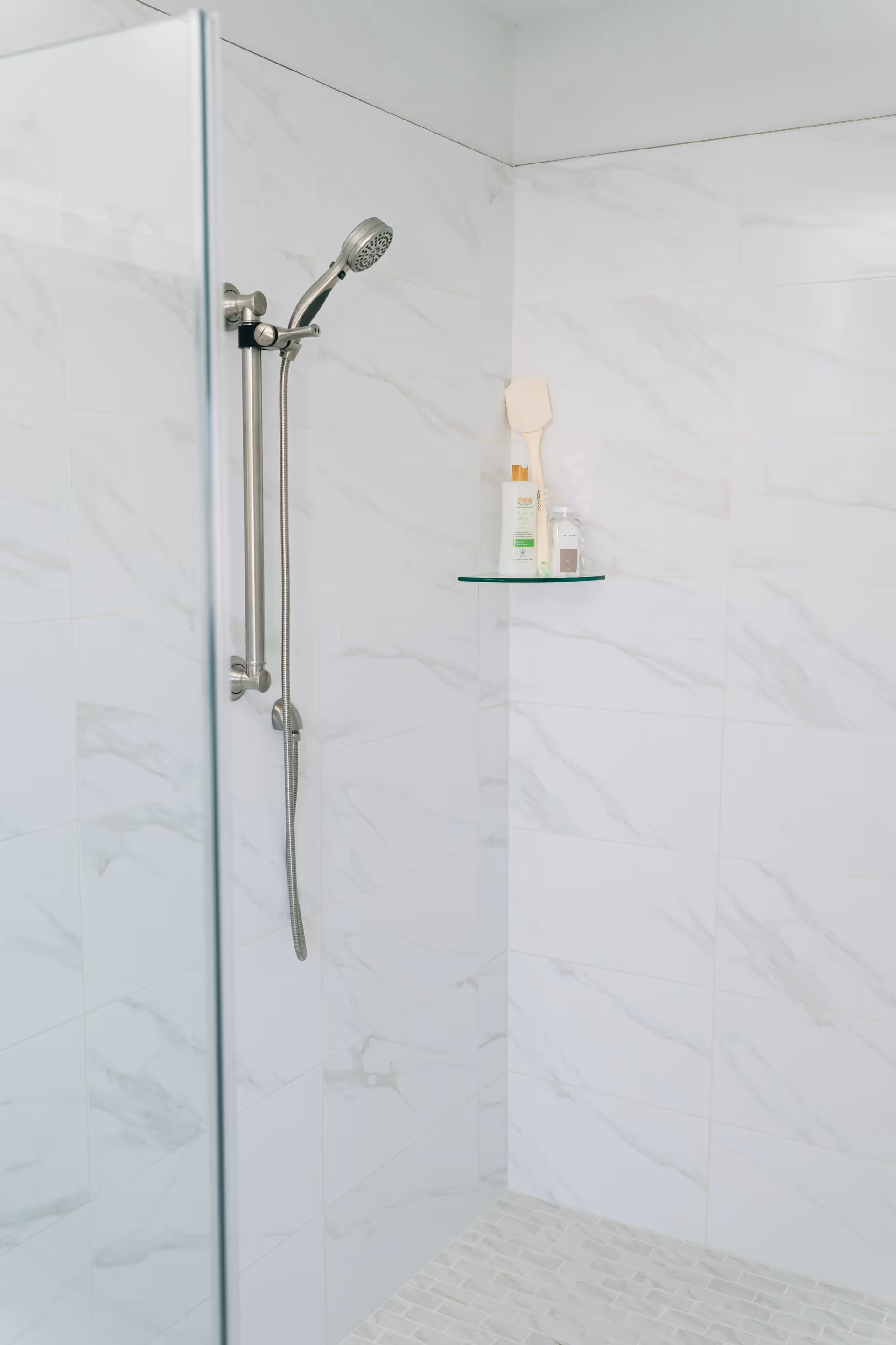 A modern shower area with white marble-patterned tiles on the walls and floor. There is a silver handheld showerhead attached to a vertical bar on the wall. A small glass shelf holds a wooden bath brush, a bottle of lotion, and two small containers.
