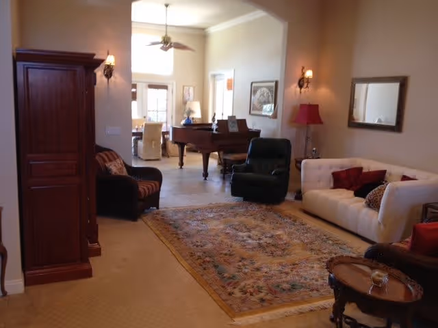A cozy living room with a white couch, a black armchair, a wooden armoire, a small round wooden table, and a patterned area rug. In the background, there is a grand piano and a dining area with chairs and a ceiling fan. The walls are light-colored with wall sconces and framed artwork.
