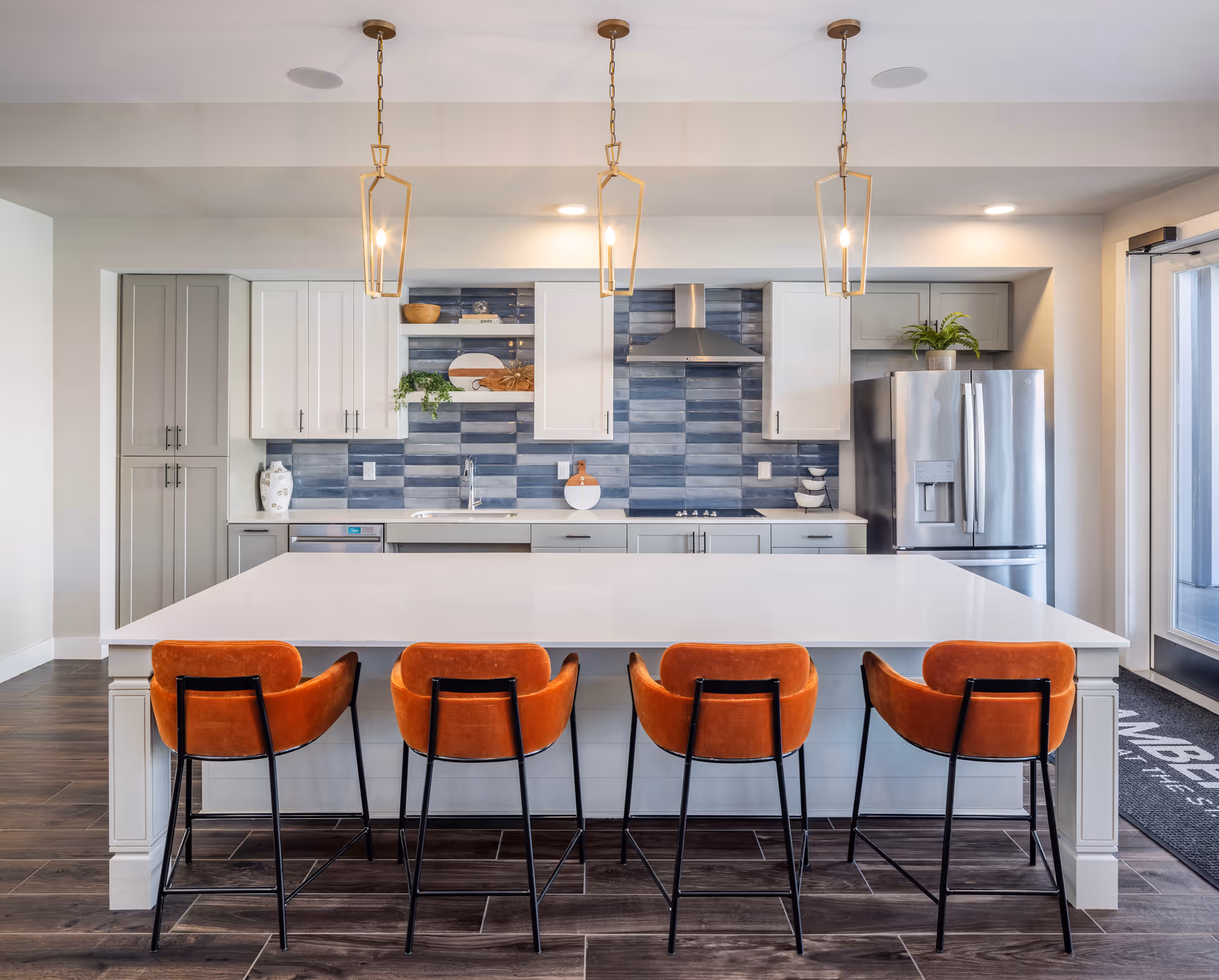 Modern kitchen with a large white island countertop and four orange bar stools. The kitchen features gray and white cabinetry, a stainless steel refrigerator, a built-in stovetop with a stainless steel range hood, and blue-gray tiled backsplash. Three pendant lights hang above the island, and there is a glass door to the right with a mat that reads 'Amberlin at The Station'.