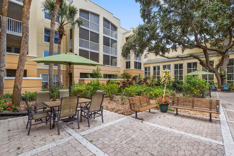 Outdoor courtyard area of a senior living facility with patio tables and chairs under green umbrellas, wooden benches, potted plants, and surrounding greenery. The building has multiple floors with screened balconies and large windows.