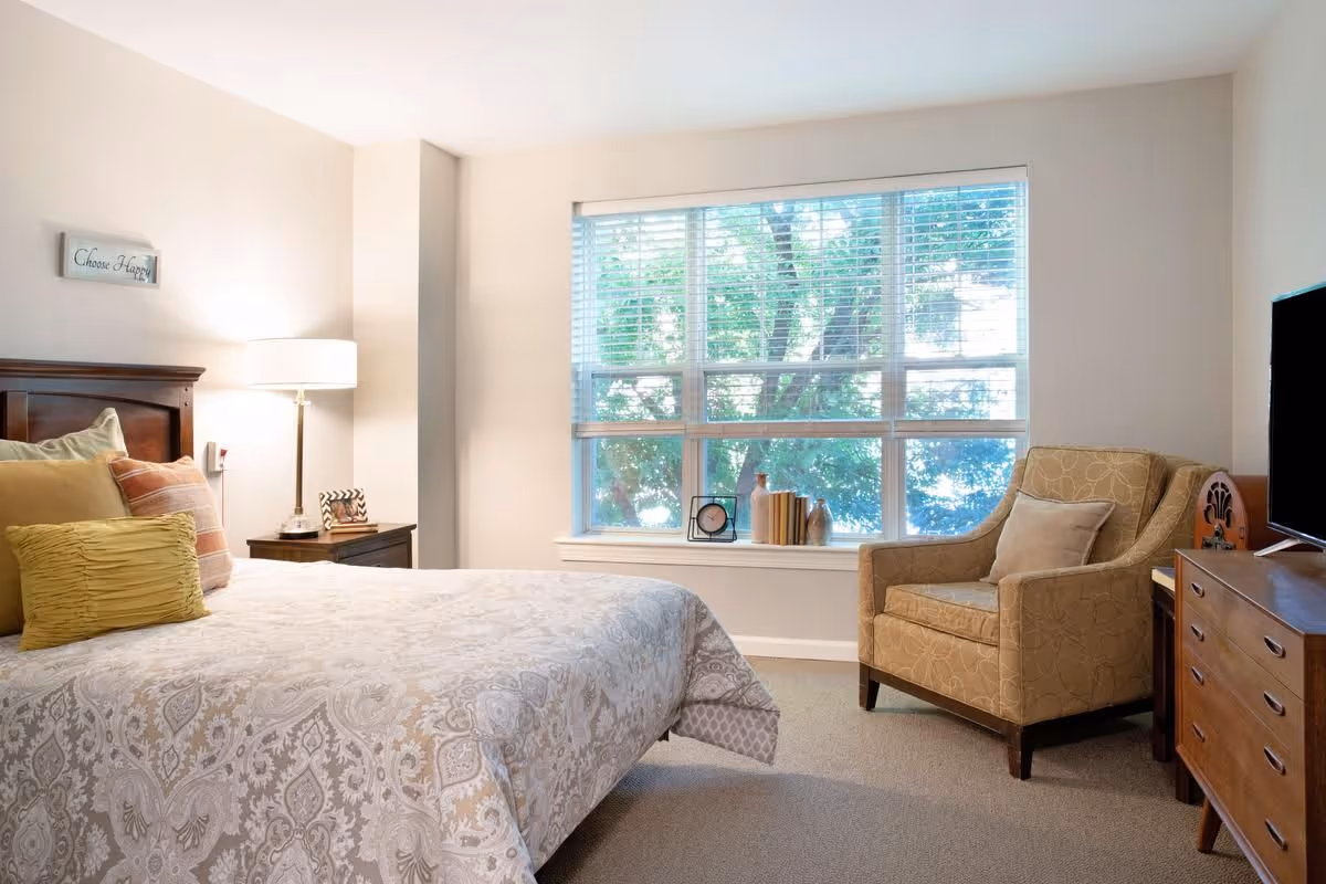 Sunlit bedroom with a bed, bedside table and lamp, an upholstered armchair, dresser, and a large window with blinds.
