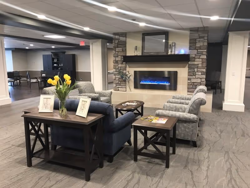 A cozy senior living common area with a modern electric fireplace set in a stone and tile surround. The room features a seating arrangement with two gray patterned armchairs, a blue armchair, and a wooden coffee table with magazines. A wooden console table behind the blue armchair holds a vase with yellow tulips and two framed pictures. The space has carpeted flooring, white pillars, and a ceiling with recessed lighting. In the background, there are additional tables and chairs and a dark wooden cabinet.