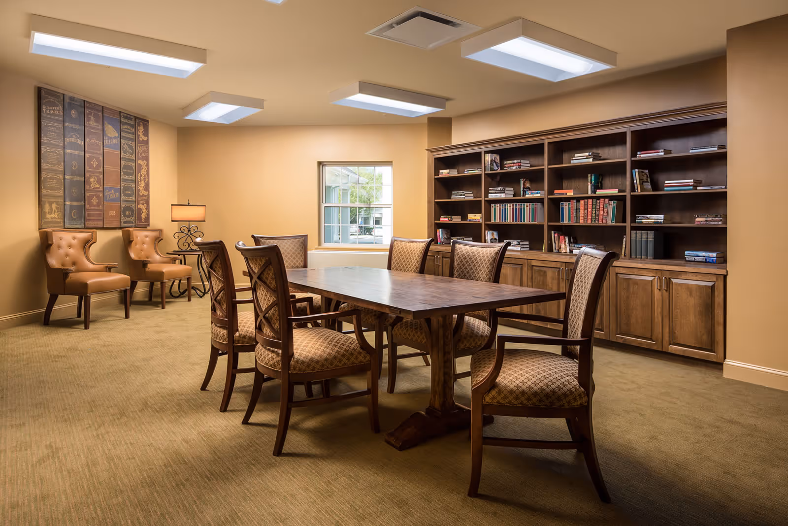 A warm interior room with a large wooden table surrounded by upholstered chairs, built-in bookshelves, and two leather armchairs by a lamp.