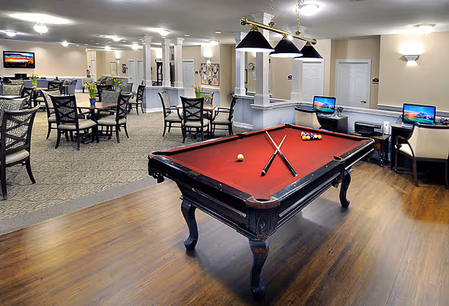A spacious senior living common area featuring a red felt pool table with two crossed cues and billiard balls set up. In the background, there are multiple round tables with chairs arranged for dining or socializing. The room has beige walls, carpeted and wooden flooring, and several computer stations with monitors on desks along the wall. Ceiling lights and wall sconces provide illumination.