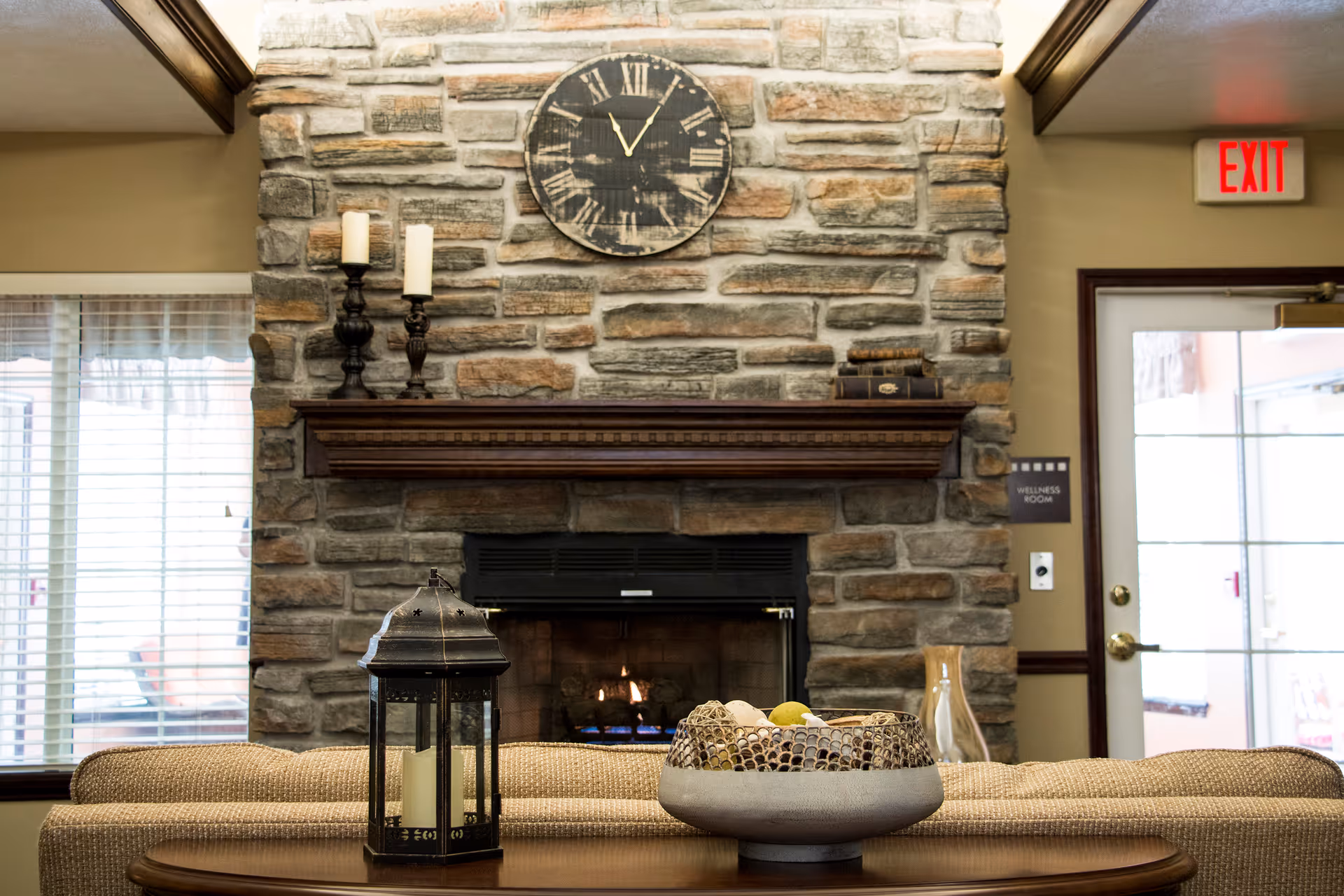 Cozy interior living area with a stone fireplace, a wooden mantel holding two candles and some books, a large round clock above the mantel, a beige couch facing the fireplace, a decorative lantern with candles on a wooden table, and a bowl of decorative items. There is a window with blinds on the left and a glass door with an exit sign and a 'Wellness Room' sign on the right.