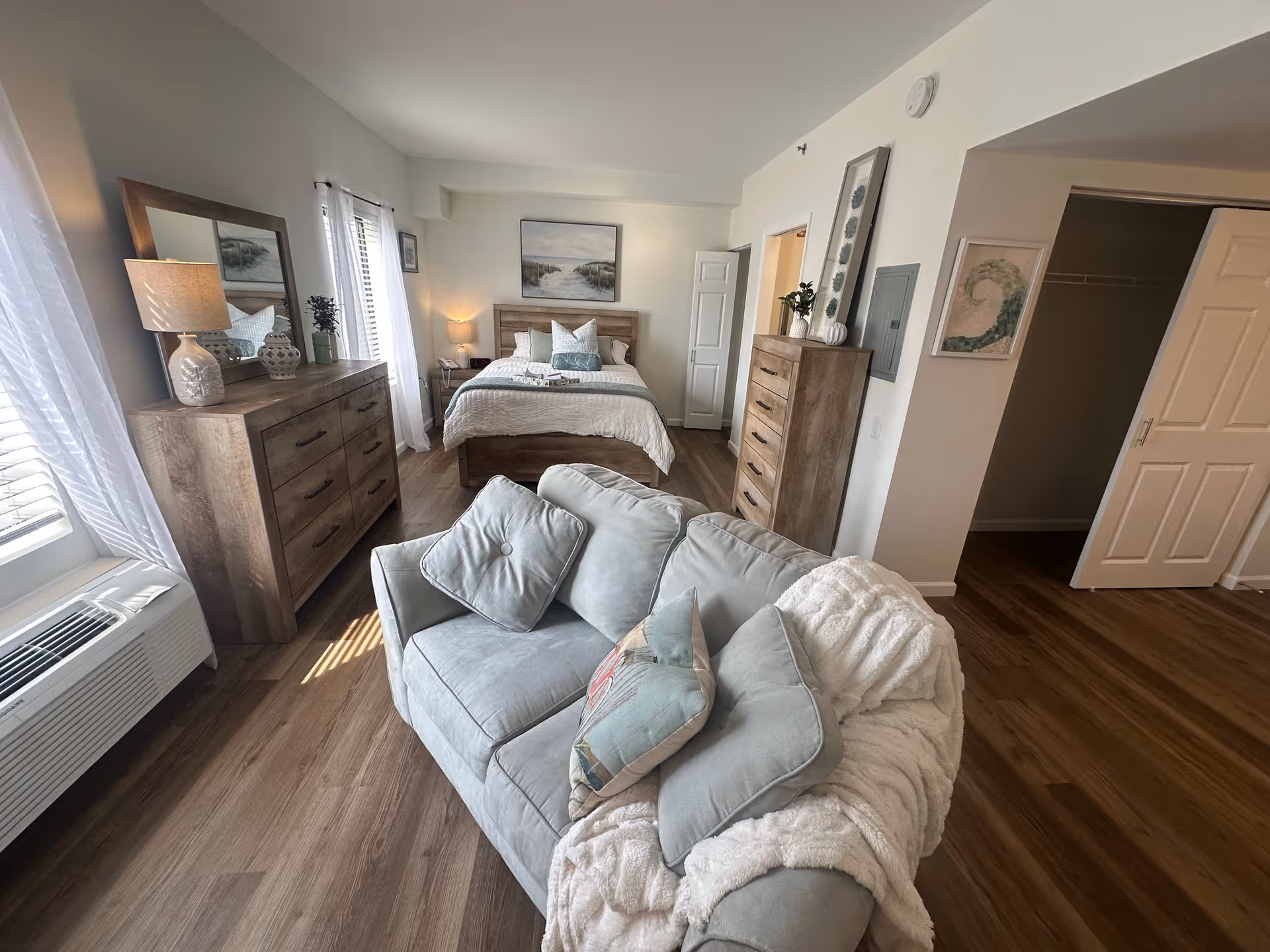 Bright furnished studio apartment showing a couch in the foreground, a bed with nightstands, and wooden dressers against light walls.