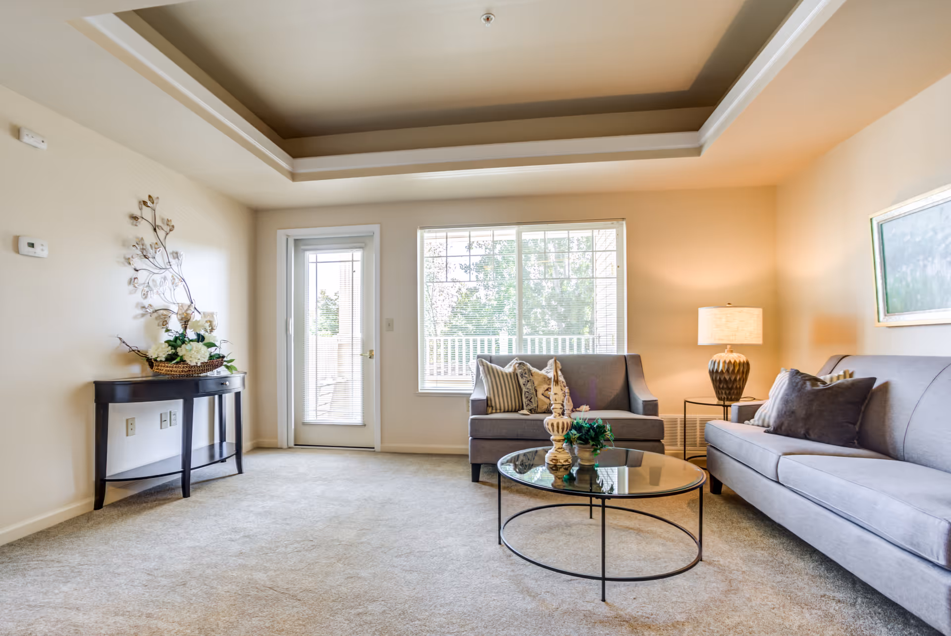 A bright living room with beige walls and carpet, featuring two gray sofas with decorative pillows, a round glass coffee table with ornaments, a side table with a lamp, a wall-mounted painting, a small black console table with a floral arrangement, and a large window and glass door leading to an outdoor area.