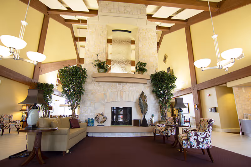 Spacious living room area with high ceilings and exposed wooden beams, featuring a large stone fireplace in the center. The room is furnished with a beige sofa, patterned armchairs, side tables with lamps, and tall green plants on either side of the fireplace. Modern hanging light fixtures illuminate the space.