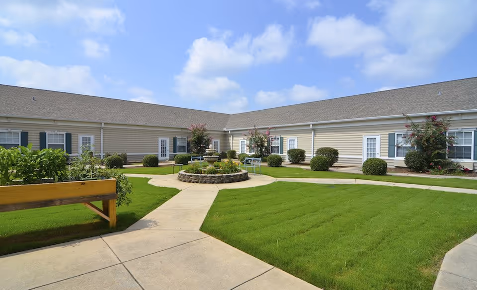 A courtyard area with green grass, a circular stone flower bed with a small fountain in the center, surrounded by benches. The courtyard is enclosed by a single-story building with beige siding, white doors, and windows with dark shutters. The sky is blue with some clouds.