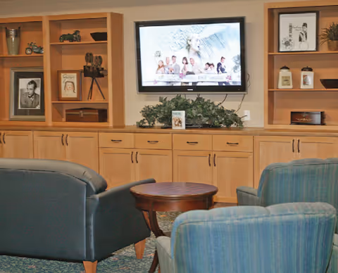 A communal seating area with upholstered chairs facing a wall-mounted TV above built-in wooden cabinets and decorative shelving.