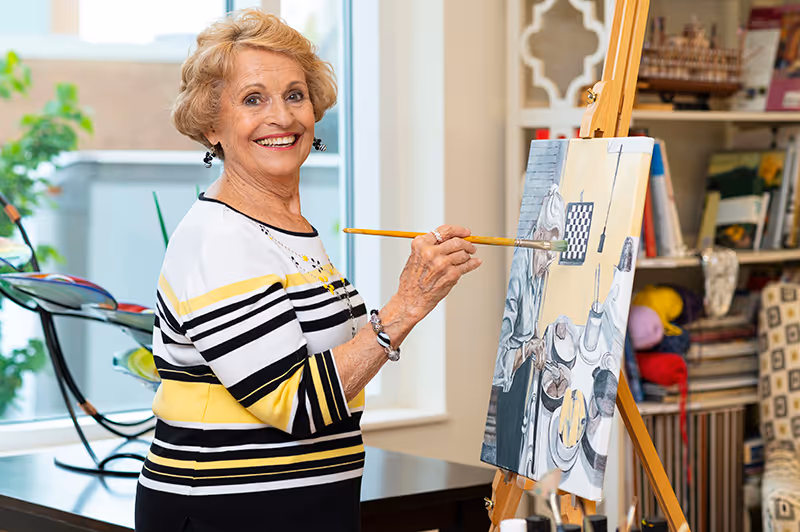 An elderly woman with short blonde hair wearing a striped black, white, and yellow top is smiling while painting on a canvas set on an easel in a well-lit room with shelves and art supplies in the background.
