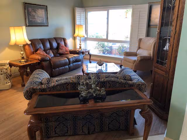 A cozy living room with a brown leather sofa, a beige armchair, a patterned upholstered loveseat, and wooden tables with lamps. There is a large window with white shutters letting in natural light, and a wooden cabinet with glass doors on the right side.