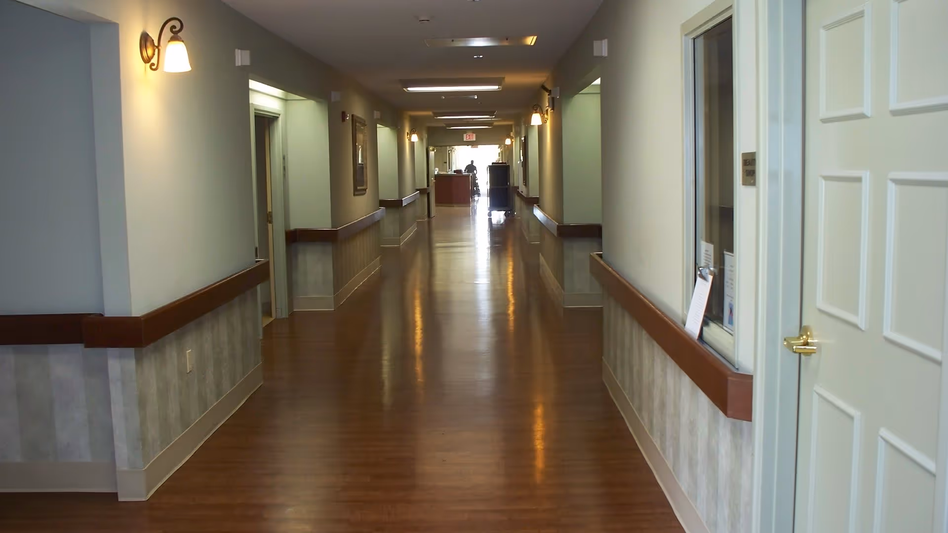 A long, well-lit hallway in a nursing and rehabilitation facility with wooden floors, beige walls, and handrails on both sides. There are wall-mounted lights and several doorways along the corridor. At the far end, a person in a wheelchair is visible near an exit door.
