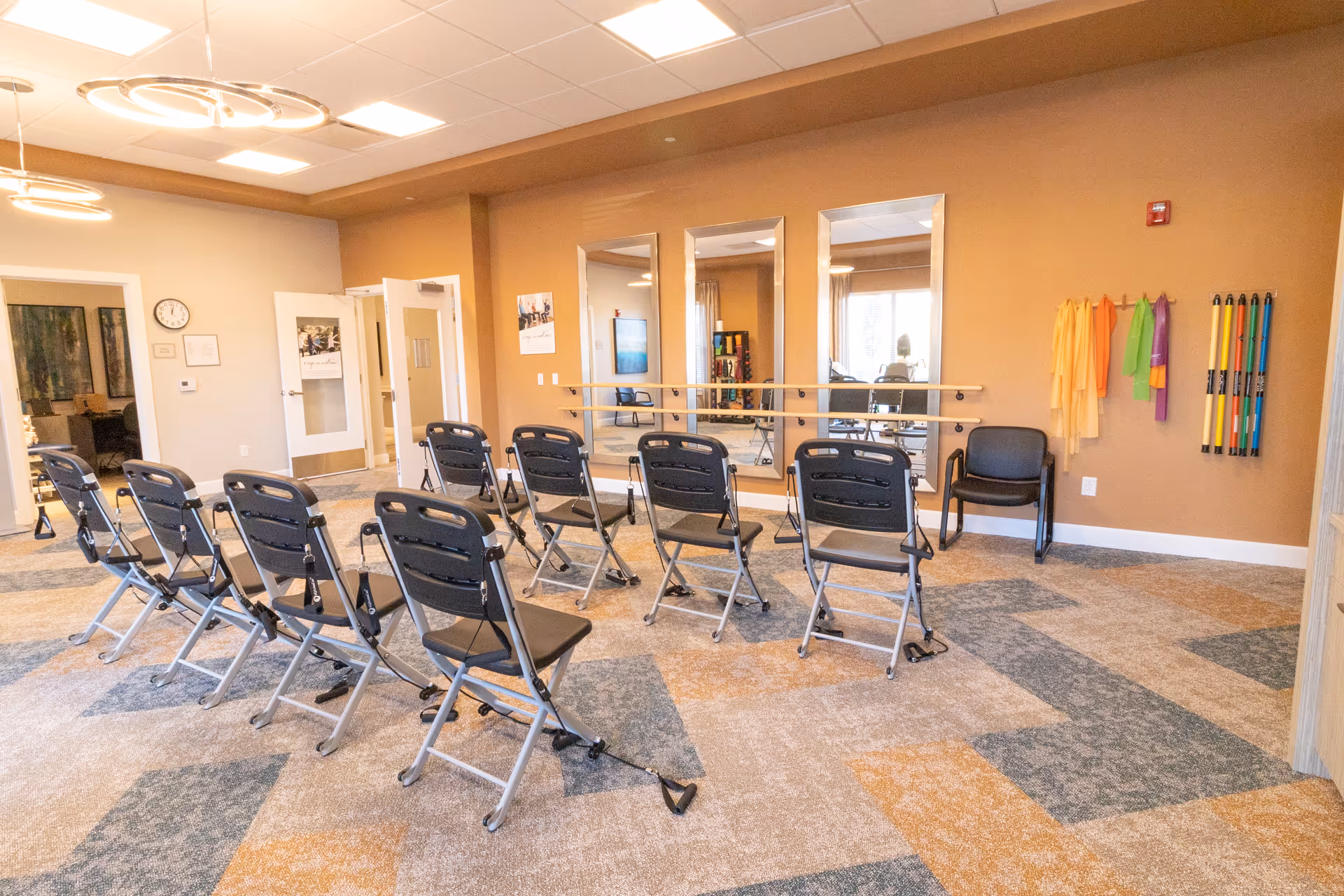 Activity/exercise room with rows of folding chairs facing wall mirrors, a wall-mounted barre, and hanging exercise bands.