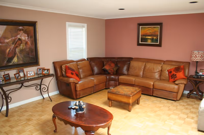 A cozy living room with a large brown leather sectional sofa adorned with red and brown decorative pillows. In front of the sofa is a wooden coffee table with a small decorative item on it. The room has parquet flooring and walls painted in two shades of pink. On the left wall, there is a framed painting of a woman playing a cello above a decorative console table with several framed photos. On the right wall, there is a framed painting of a sunset over water and a side table with a patterned lamp and some books.