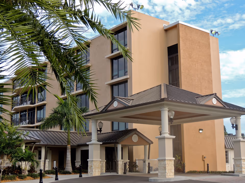 Exterior view of a multi-story beige building with large windows and a covered entrance supported by white columns. Palm trees and other greenery are visible in the foreground under a partly cloudy sky.