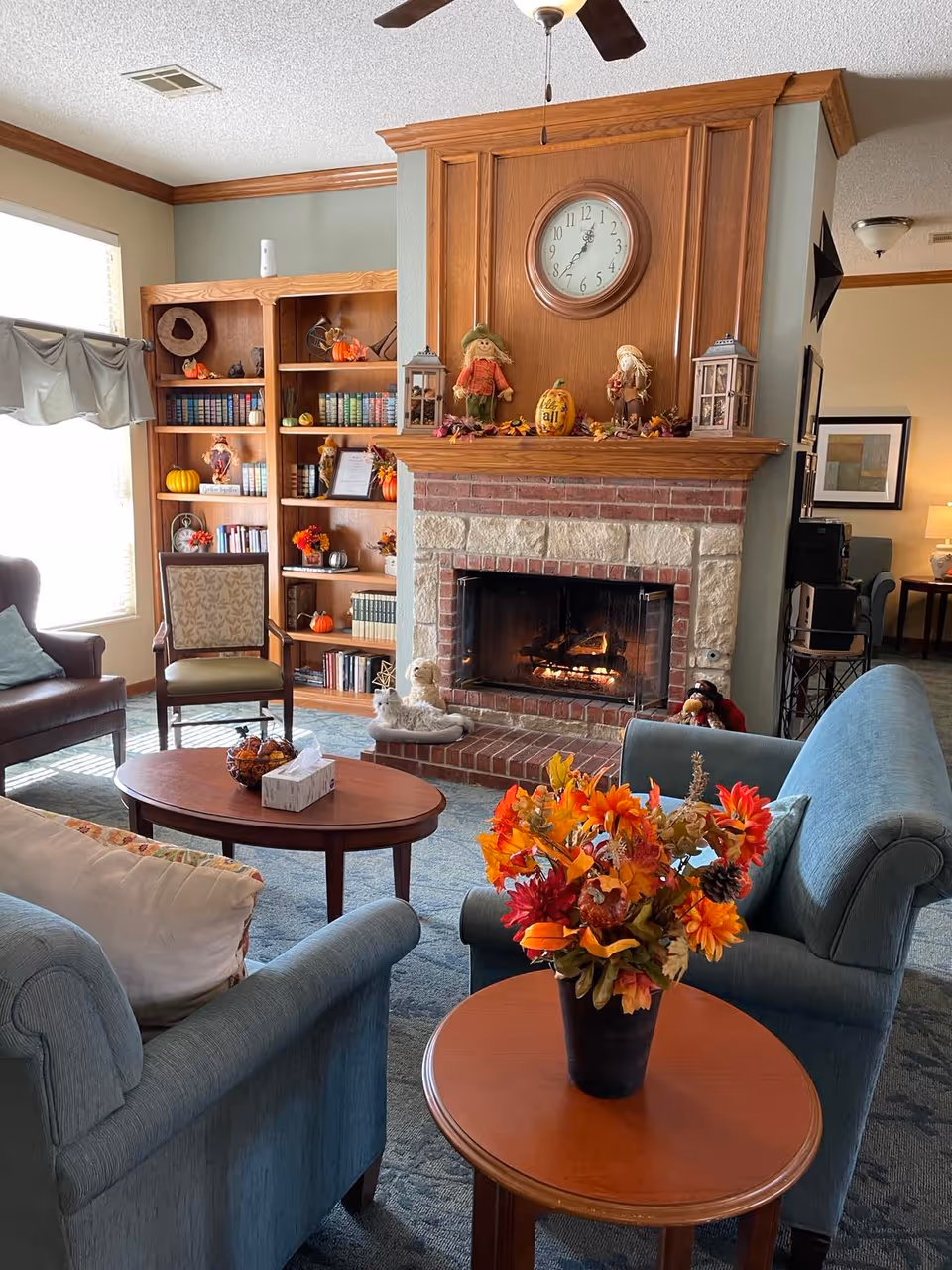 A cozy living room with a lit brick fireplace decorated with autumn-themed items including scarecrows and a pumpkin. Above the fireplace is a round clock. The room features blue upholstered chairs, a wooden coffee table with a tissue box and decorative bowl, and a side table with a vase of vibrant fall flowers. A wooden bookshelf filled with books and small decorations is visible in the background, along with a window letting in natural light.