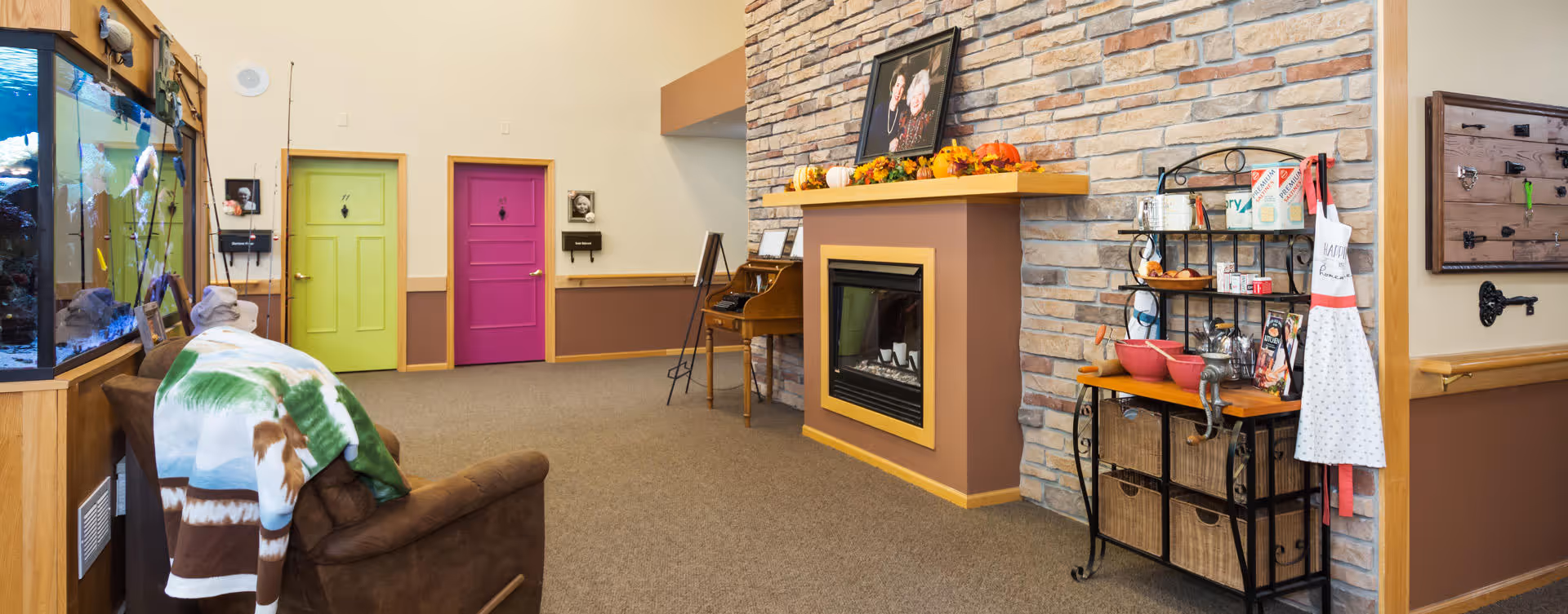 Interior view of a cozy assisted living facility common area featuring a brown armchair with a colorful blanket draped over it, a large fish tank on the left, two brightly colored doors (one green and one purple) in the background, a stone fireplace decorated with autumn-themed items and a framed photo, a small wooden desk with a chair, and a black metal shelving unit with baskets, bowls, and kitchen items including an apron hanging on the side.