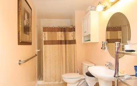 A small bathroom with beige walls featuring a shower with a beige and brown patterned curtain, a white toilet, a white pedestal sink, an oval mirror above the sink, a wall-mounted cabinet, a towel bar, and a framed picture on the wall.