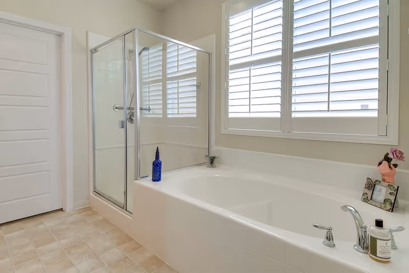 A clean and bright bathroom featuring a large white bathtub with a silver faucet, a glass-enclosed shower with a silver frame, a window with white plantation shutters, and a tiled floor. There are decorative items including a blue bottle on the bathtub edge, a small plant, a framed picture, and a pink flower vase.