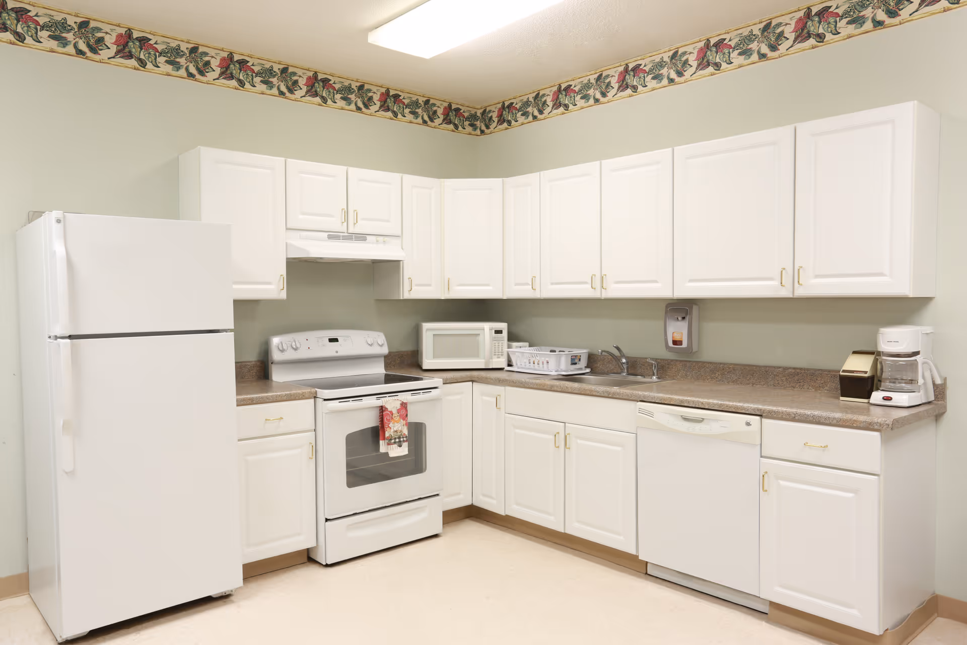 A clean and well-lit kitchen with white cabinets and appliances including a refrigerator, stove, microwave, dishwasher, and coffee maker. The walls are light green with a floral border near the ceiling.