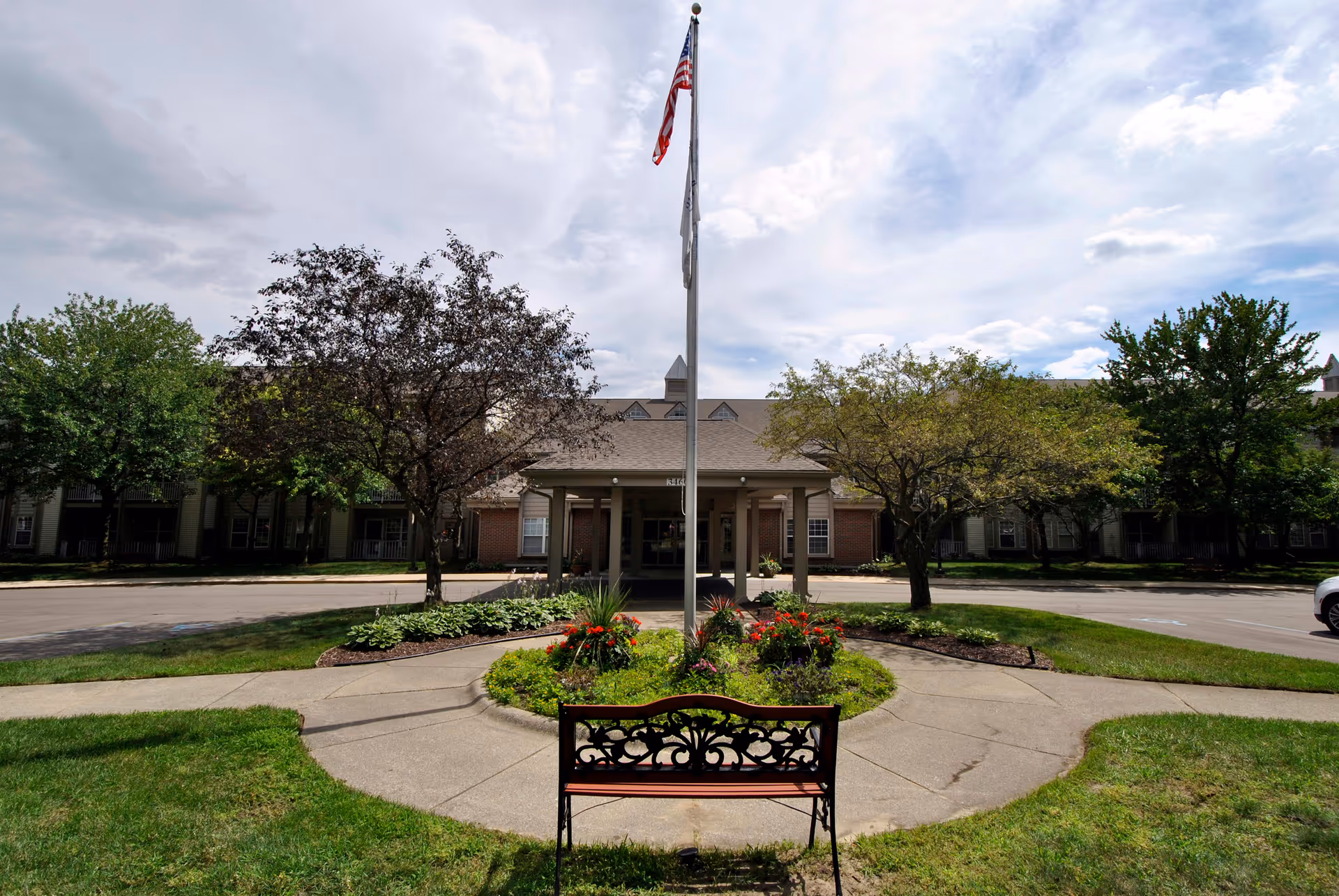 Front exterior view of Westhaven Manor facility with a circular driveway, a flagpole with an American flag, a garden with flowers, trees, and a decorative bench in the foreground under a partly cloudy sky.