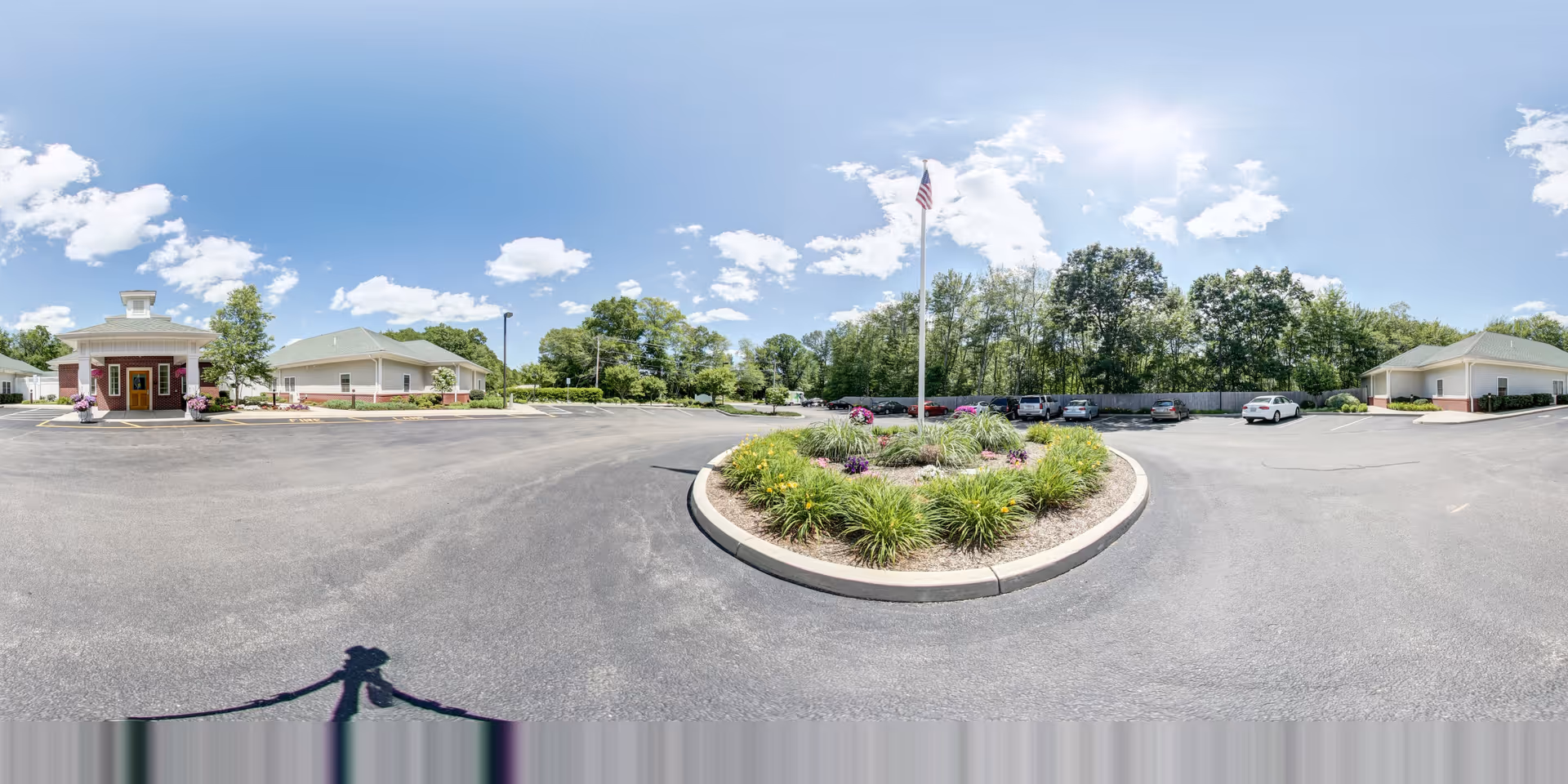 Wide panoramic view of the parking lot and entrance area of The Lighthouse at Lincoln senior living facility on a sunny day with blue sky and scattered clouds. The image shows a circular landscaped island with flowers and an American flag in the center of the parking lot. The facility buildings are visible on the left and right sides, surrounded by trees and greenery.