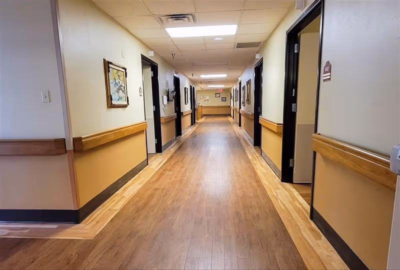 A long, well-lit hallway in a nursing and rehabilitation facility with wooden flooring and beige walls. Several doorways line both sides of the corridor, some with room number signs. Handrails run along the walls, and framed pictures are hung at intervals.
