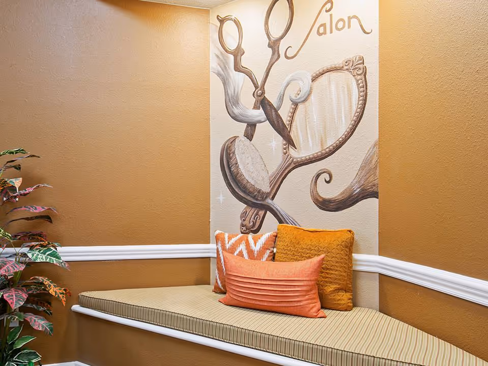 A cozy corner seating area with a cushioned bench and three decorative pillows in orange and patterned designs. The wall behind features a mural with large illustrations of hair salon tools including scissors, a hairbrush, and a hand mirror, with the word 'Salon' written above. The walls are painted in warm brown tones with white trim, and there is a leafy plant to the left.