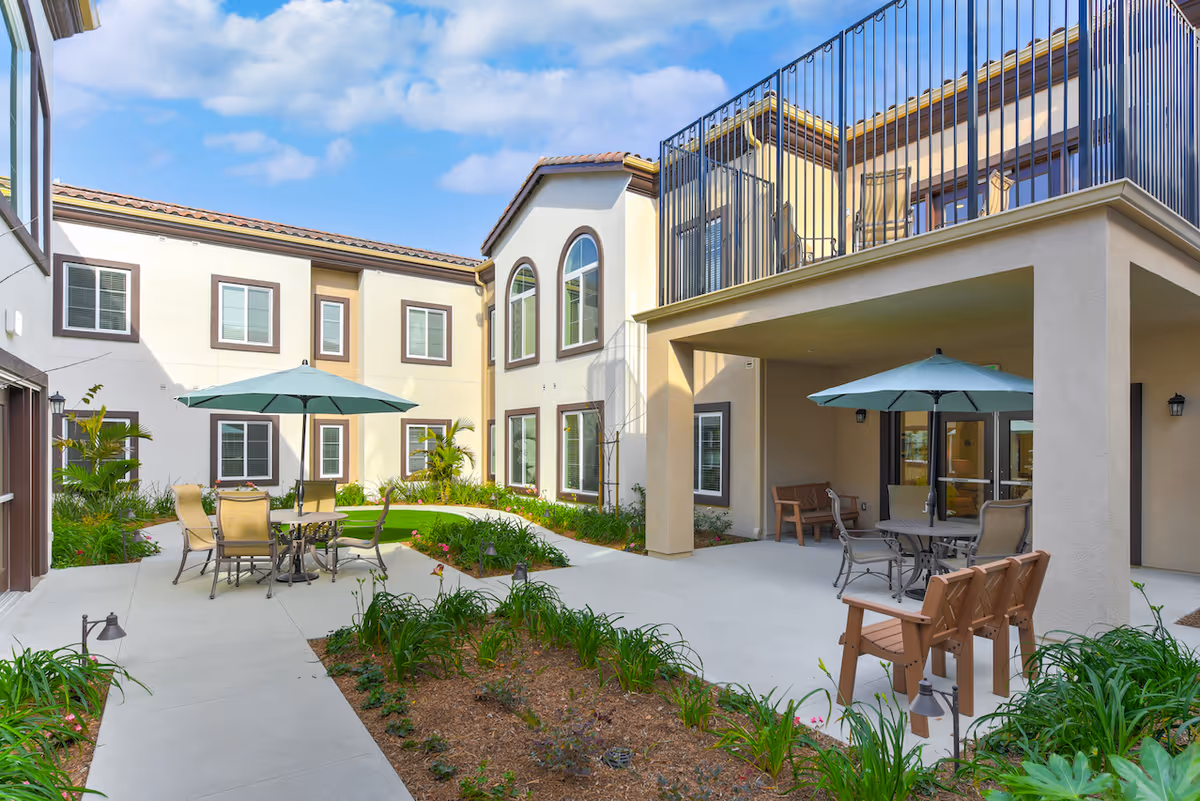 Sunny two-story courtyard at Silverado Thousand Oaks with patio tables, umbrellas, chairs, planted beds and walkways surrounded by the building.