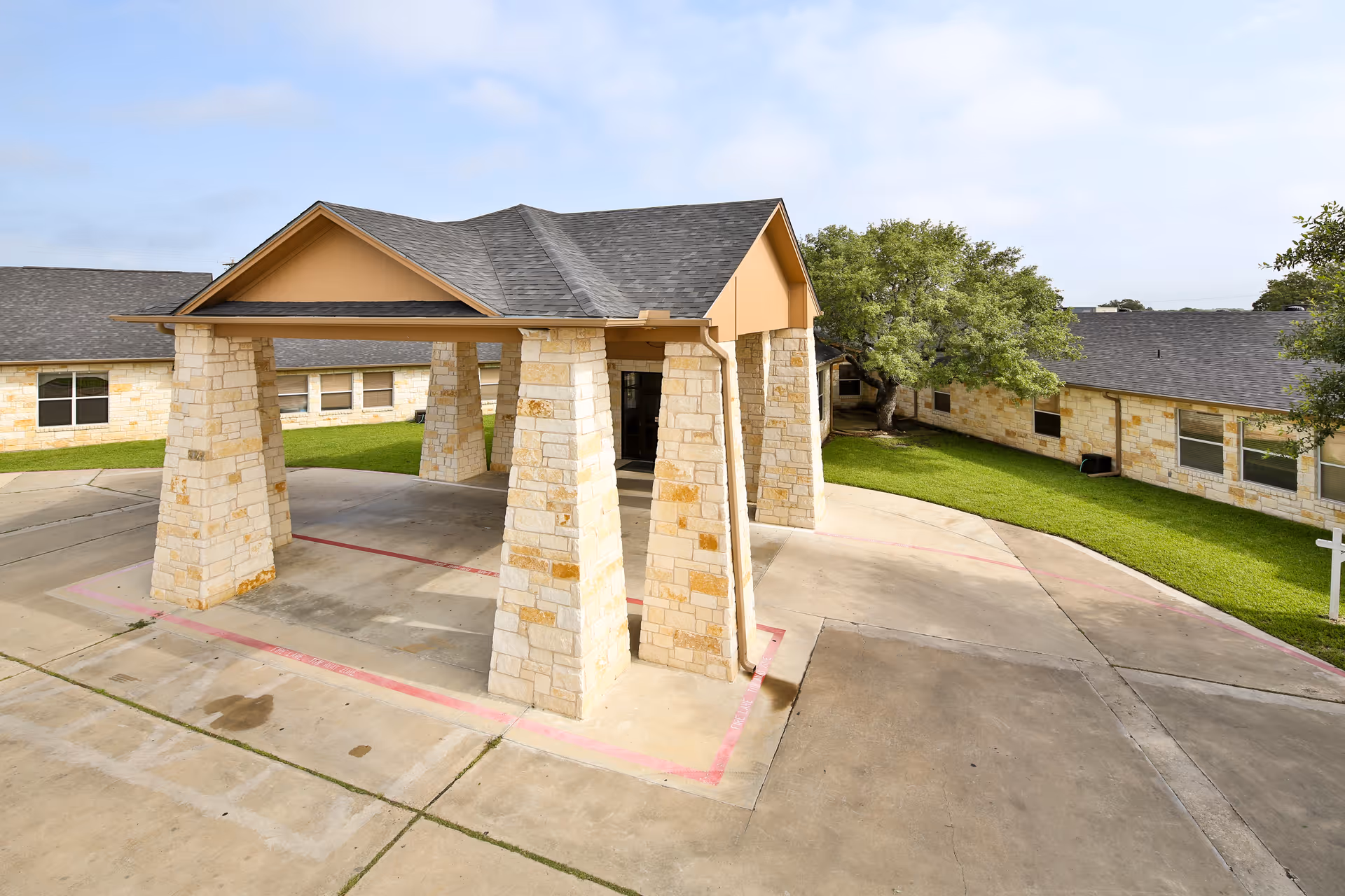 Drive-through covered entrance with stone pillars at the front of a low stone-faced nursing facility surrounded by lawn and pavement.