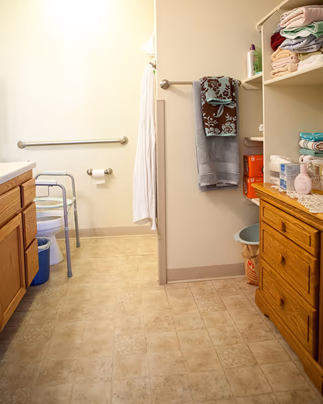 A bathroom with beige tiled floor and light-colored walls. There is a wooden cabinet with drawers on the right side and shelves above it holding folded towels and toiletries. A towel rack on the wall holds two towels, one gray and one brown with a floral pattern. On the left side, there is a toilet with a metal grab bar and a walker nearby. A white shower curtain is partially visible in the background.