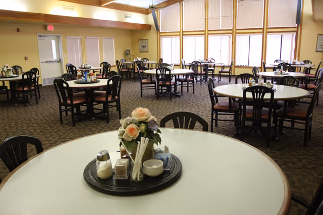 A spacious dining room with multiple round tables and black chairs. Each table has a centerpiece with flowers and condiments. Large windows with vertical blinds allow natural light to fill the room. The carpet is patterned, and there is a door and framed pictures on the walls.