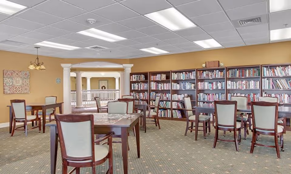 Bright communal library/activity room with bookshelves along the wall, several tables and upholstered chairs, and decorative columns opening to an interior railing.