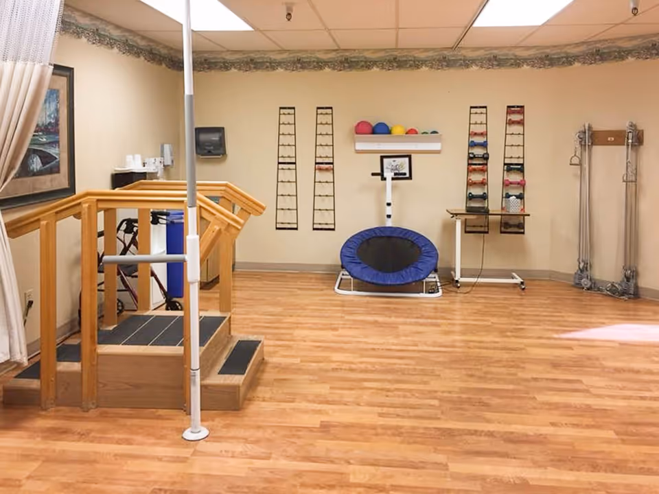 Rehabilitation therapy room with wooden parallel bars, a small exercise trampoline, medicine balls and dumbbells on racks.