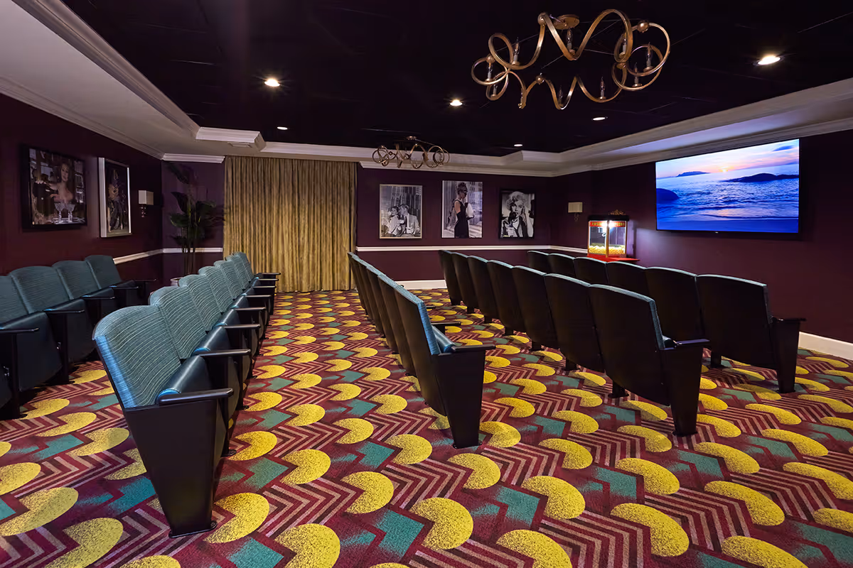A small theater room with rows of green cushioned seats facing a large screen displaying a sunset over the ocean. The room has a colorful patterned carpet, dark purple walls adorned with black and white celebrity photos, and a popcorn machine near the screen. Two decorative chandeliers hang from the ceiling.