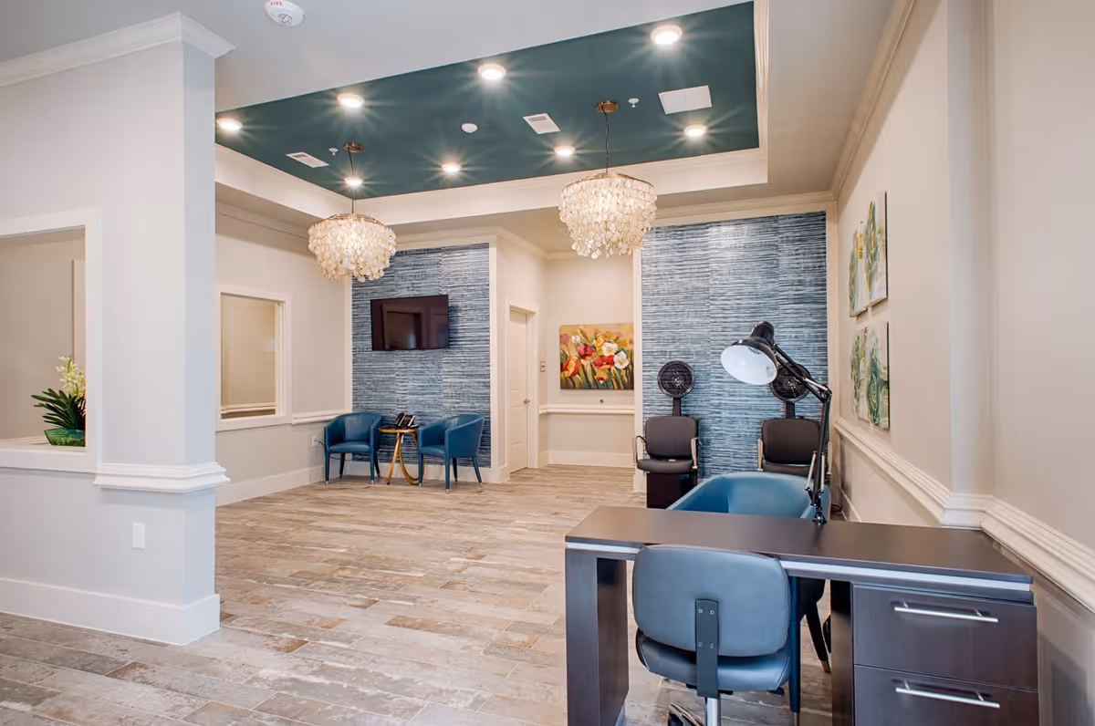 A modern and spacious interior room with a desk and chair in the foreground, two black chairs with a lamp behind the desk, and two blue chairs with a small table in front of a blue textured accent wall with a mounted TV. The ceiling has recessed lighting and two decorative chandeliers, and there are paintings on the walls.