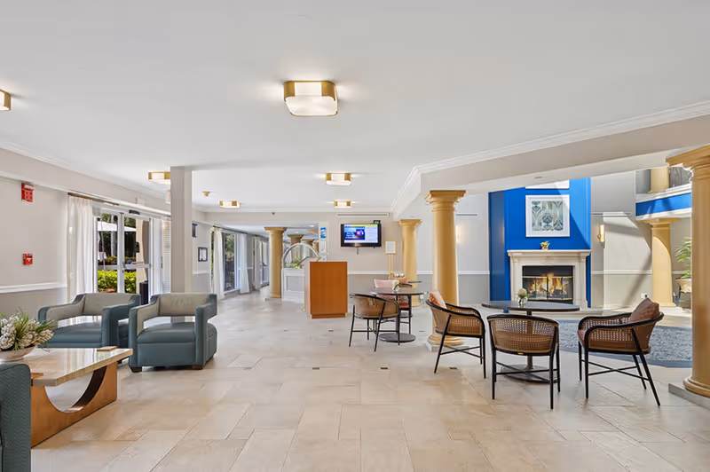 Spacious and well-lit common area in a senior living facility featuring comfortable seating with armchairs and small tables, a fireplace with a blue accent wall, large windows with sheer curtains allowing natural light, and decorative columns.