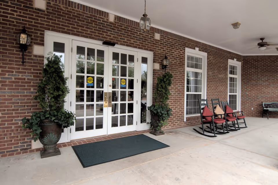 Covered entrance area of a building with double glass doors framed in white, flanked by two large potted plants. The brick exterior wall has two windows with white trim, and there are three black rocking chairs with red and beige cushions arranged beside the windows. A ceiling fan and light fixtures are mounted on the white ceiling above the entrance.