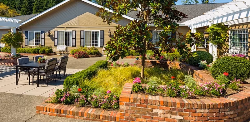 Outdoor patio area at Canterbury Gardens Memory Care Community featuring a brick planter with green shrubs and colorful flowers, a small tree in the center, a table with six chairs on a paved surface, and beige buildings with white pergolas and windows in the background under a clear blue sky.