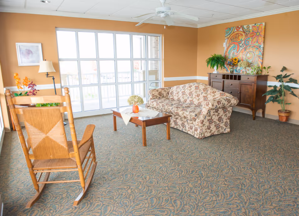 A bright living room with a floral patterned sofa, wooden coffee table with a vase of flowers, wooden rocking chair, a dark wooden sideboard with plants and colorful artwork above it, and large windows letting in natural light.